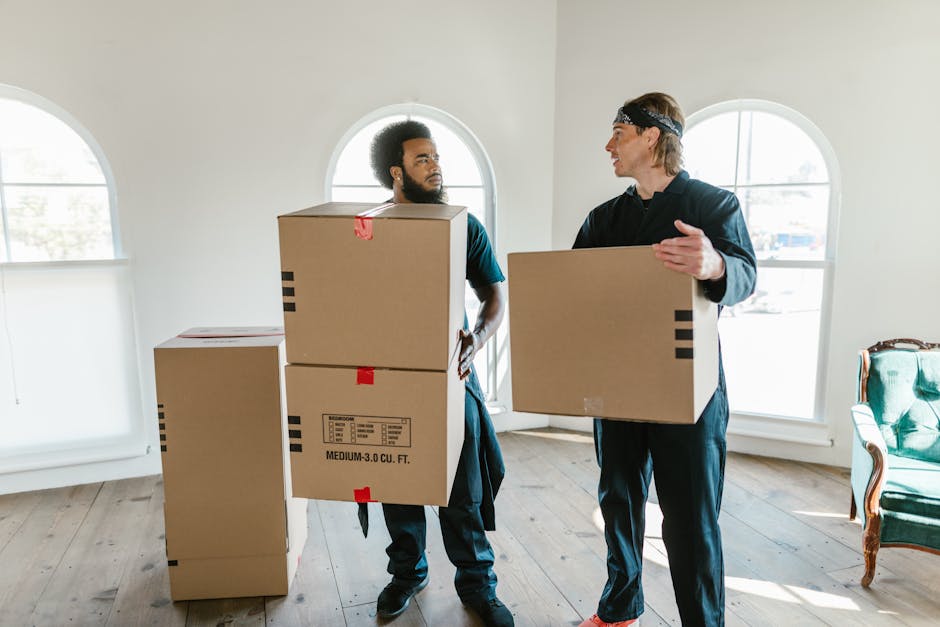 A man and a woman, both smiling gently, are standing inside a residential property with sloped ceilings and a white wall, holding large cardboard boxes filled with household items, likely during a hom