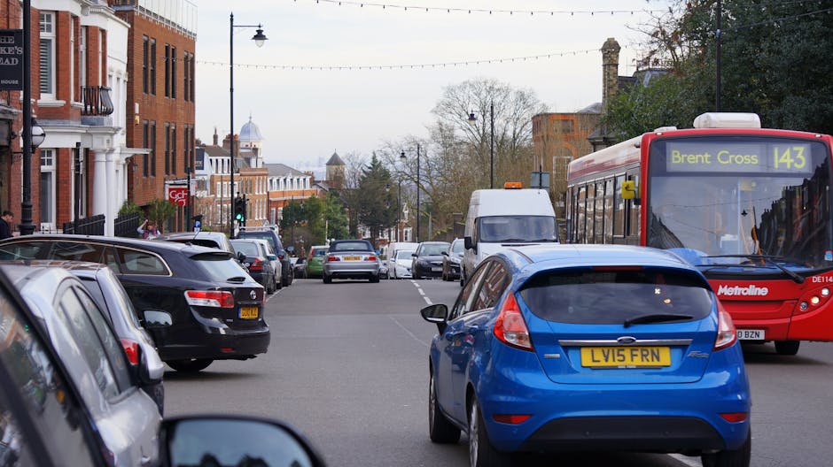 A busy residential street scene in Willesden with parked cars lining both sides of the road and moving vehicles, including a red Metroline bus on route 143 with destination 'Brent Cross' visible on th