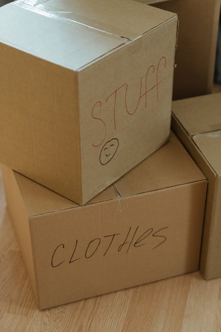 Close-up view of three cardboard boxes used for home relocation, placed on a wooden floor inside a property. The top box has the word 'STUFF' written in red marker along with a simple smiley face, indicating personal belongings contents, while the bottom box is labeled 'CLOTHES' in black marker, specifying the packed items. The boxes are standard moving cartons, with visible flaps folded and sealed for transport. In the background, additional boxes are stacked, possibly prepared for a furniture transport or packing process, reflecting an organized packing stage. The scene captures the interior environment during a packing or loading phase, with natural lighting highlighting the cardboard textures and handwritten labels, relevant to moving services provided by Man and a Van Brent, aligned with the topic of house removals and smooth moving logistics.