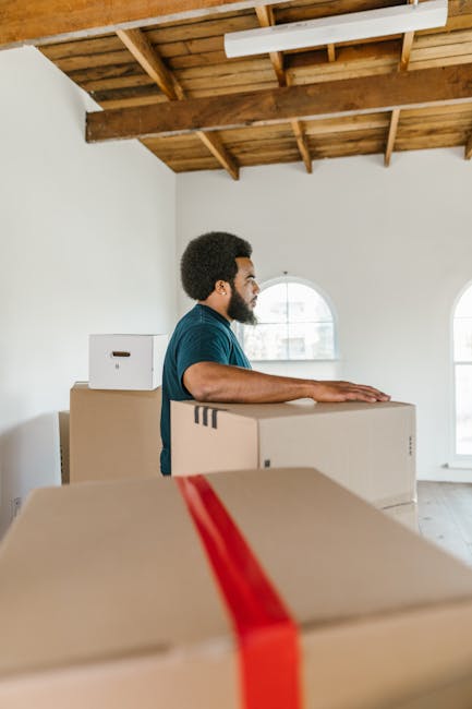 A man with a beard and curly hair wearing a blue shirt is standing inside a bright, spacious room with a high wooden ceiling and white walls. He is positioned behind several large cardboard boxes, some of which are sealed with red packing tape, indicating they are prepared for a home relocation. One of the boxes is directly in front of him, and he rests his arms on another box, suggesting he is in the process of moving or organizing belongings. The room features two arched windows, allowing natural daylight to illuminate the space, and the floor appears to be wooden. In the background, a second smaller cardboard box is visible on top of a larger one, and the overall environment suggests an ongoing packing and loading process typical of professional house removals. The company, Man and a Van Brent, may be involved in the furniture transport or packing and moving services associated with local house removals in the Willesden Green area.