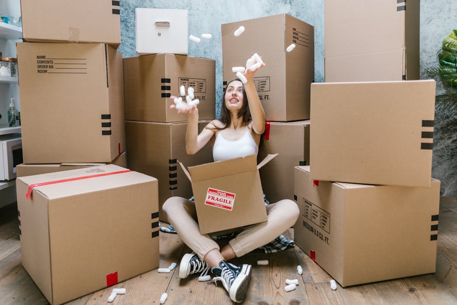 Close-up view of three cardboard boxes used for home relocation, placed on a wooden floor inside a property. The top box has the word 'STUFF' written in red marker along with a simple smiley face, indicating personal belongings contents, while the bottom box is labeled 'CLOTHES' in black marker, specifying the packed items. The boxes are standard moving cartons, with visible flaps folded and sealed for transport. In the background, additional boxes are stacked, possibly prepared for a furniture transport or packing process, reflecting an organized packing stage. The scene captures the interior environment during a packing or loading phase, with natural lighting highlighting the cardboard textures and handwritten labels, relevant to moving services provided by Man and a Van Brent, aligned with the topic of house removals and smooth moving logistics.